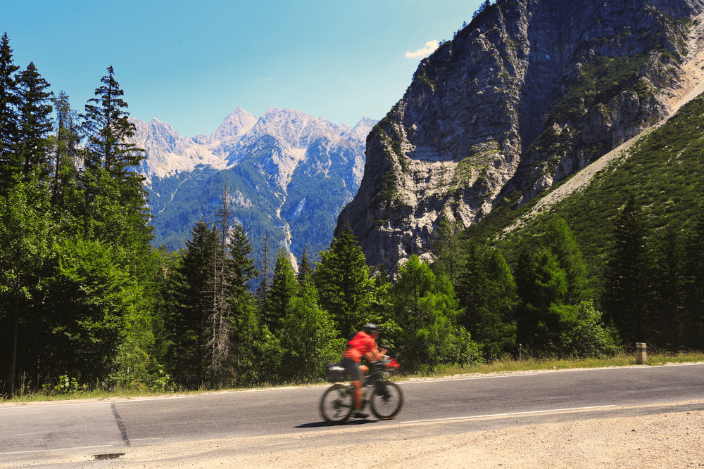 A cyclist crossing a mountain road with the Julian Alps behind them at Vršič Pass